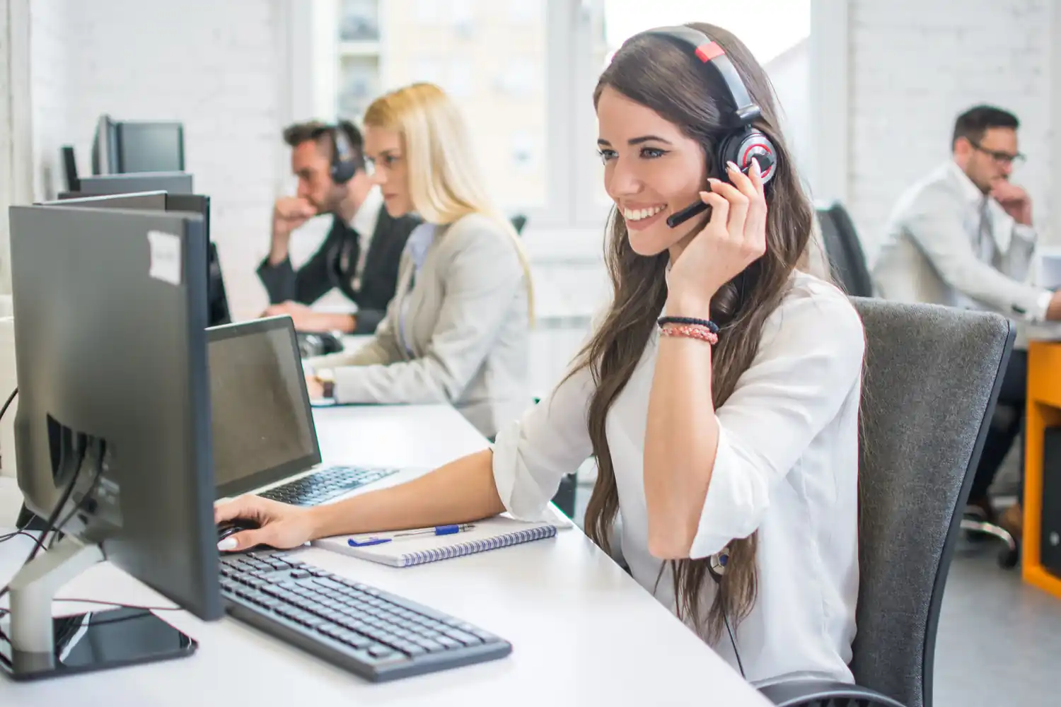 Operatrice di call center con cuffie lavora al computer in ufficio luminoso con altri colleghi.
