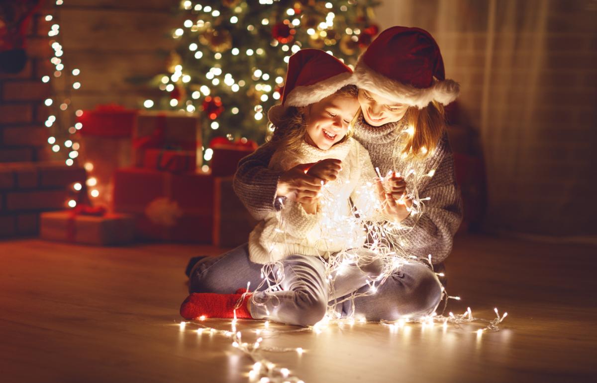 Due bambini con cappelli di Natale giocano con luci natalizie accese vicino a un albero addobbato.