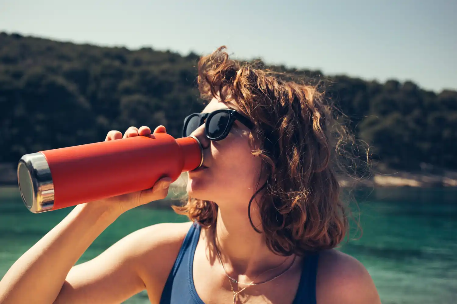 Donna beve da una borraccia rossa in una giornata soleggiata vicino a un lago.