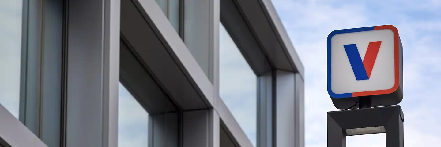 Insegna aziendale con logo V accanto a edificio moderno con vetrate, cielo azzurro sullo sfondo.