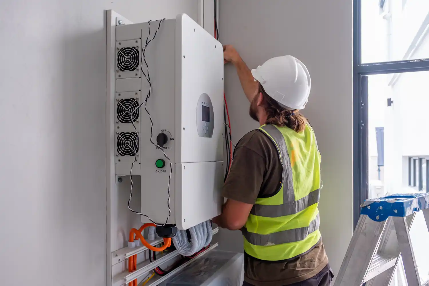 Tecnico impianta inverter su parete interna, indossa casco e gilet alta visibilità. Scala alluminio accanto.