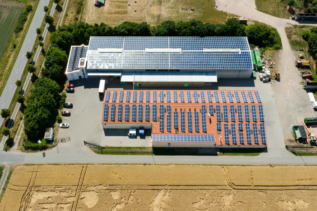Vista aerea capannone industriale con pannelli solari sul tetto, parcheggio auto e campi circostanti.