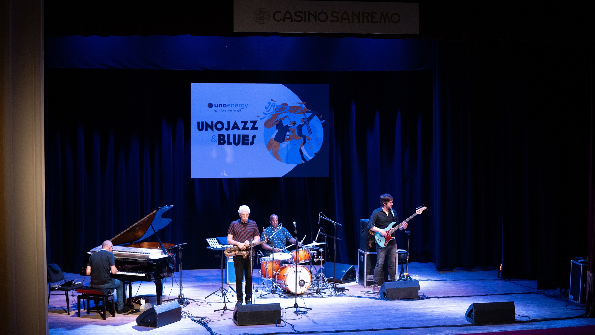 Concerto jazz in un teatro con musicisti sul palco, sfondo nero e logo Unojazz & Blues.