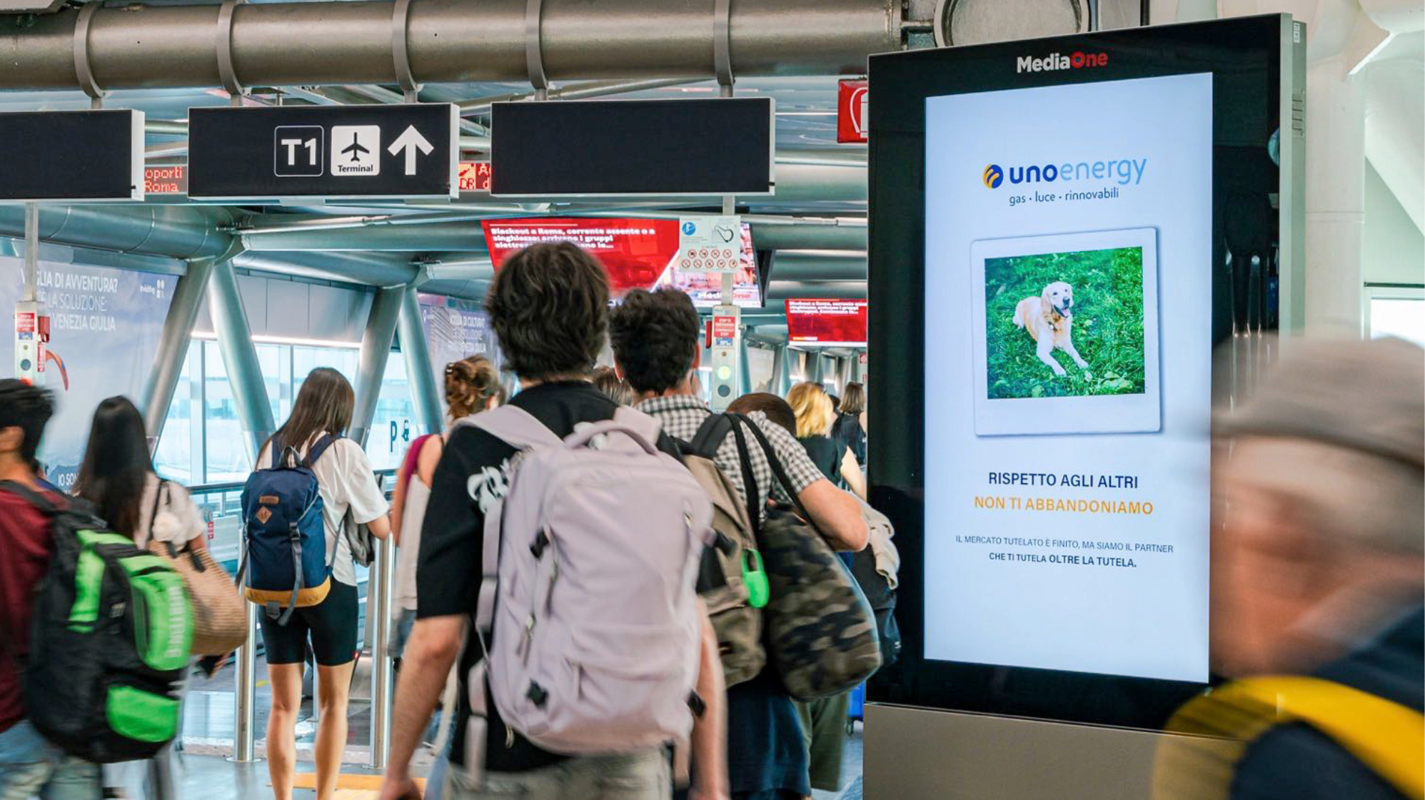 Persone camminano in aeroporto, schermo pubblicitario di Unoenergy con foto di cane e messaggio contro l'abbandono.