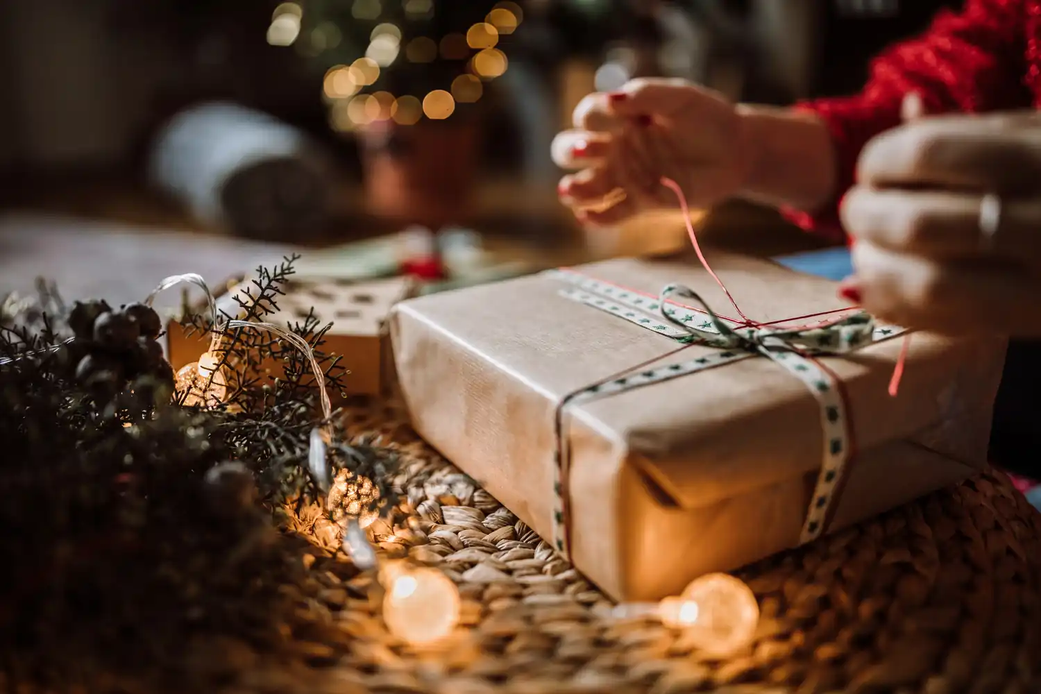 Mani legano nastro su regalo incartato, decorazioni natalizie e luci soffuse.