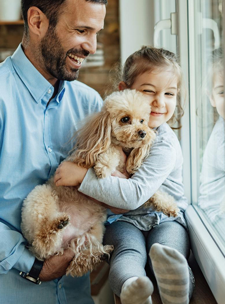 Padre e figlia sorridenti con cane in braccio, seduti vicino a una finestra.