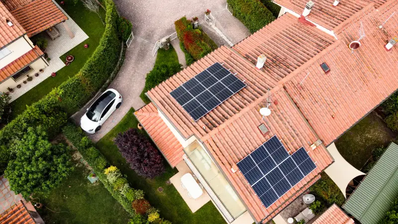 Tetto di casa con pannelli solari e auto bianca parcheggiata accanto, circondati da giardino verde.