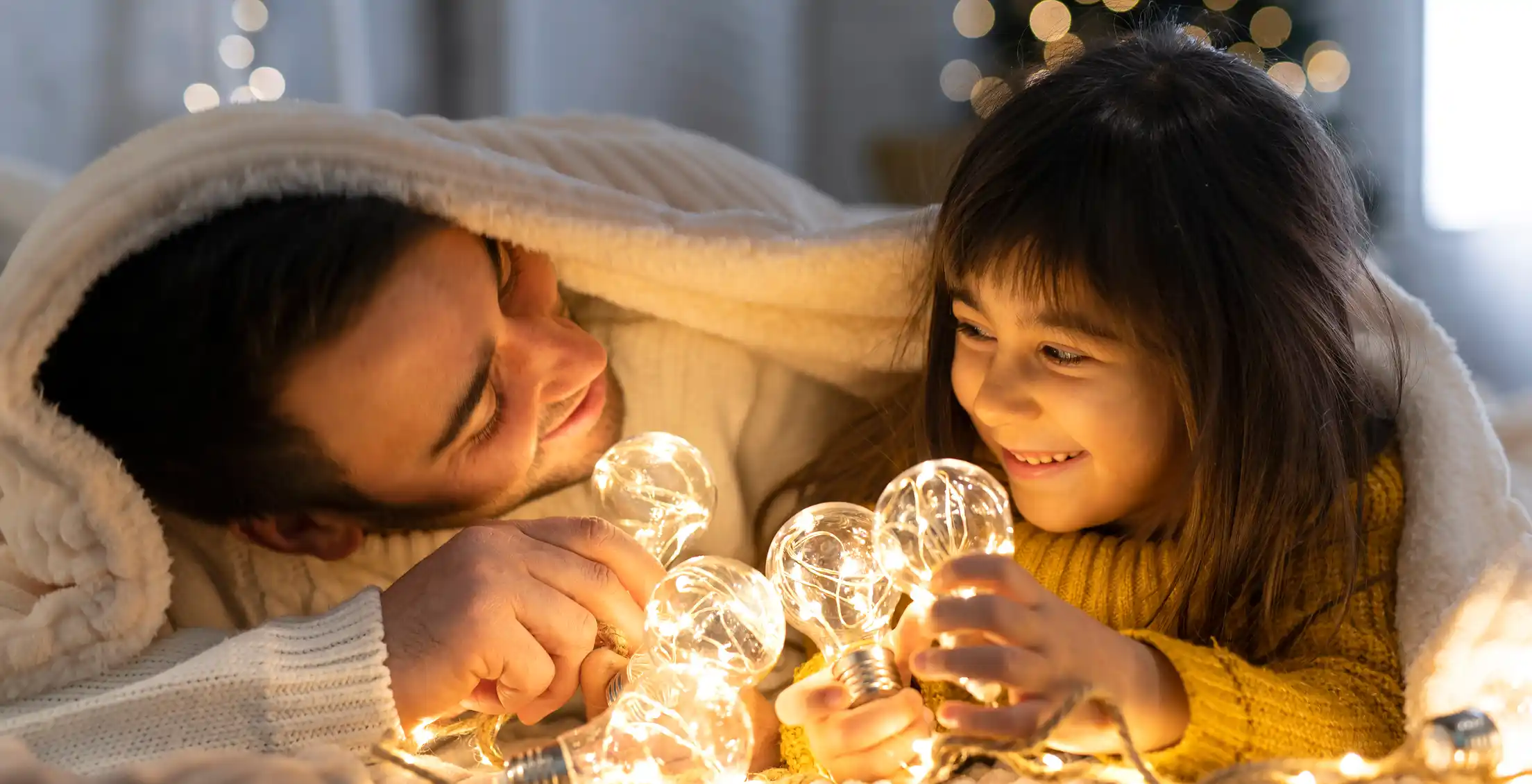 Padre e figlia sorridono sotto una coperta, giocando con luci decorative a forma di lampadina.