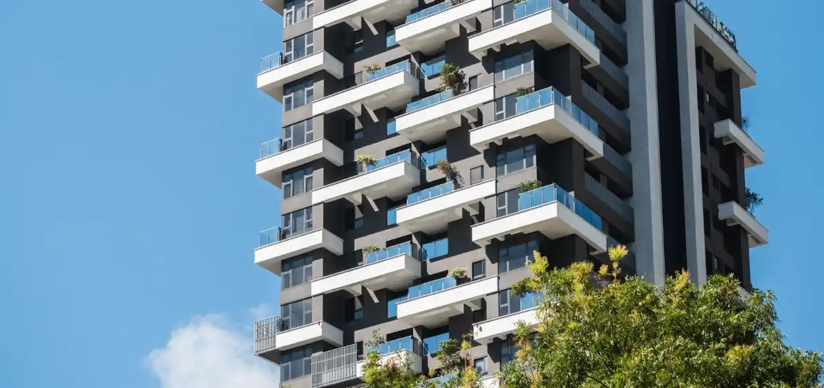 Grattacielo moderno con balconi a cielo aperto, piantumato e alberi in primo piano, cielo azzurro sullo sfondo.
