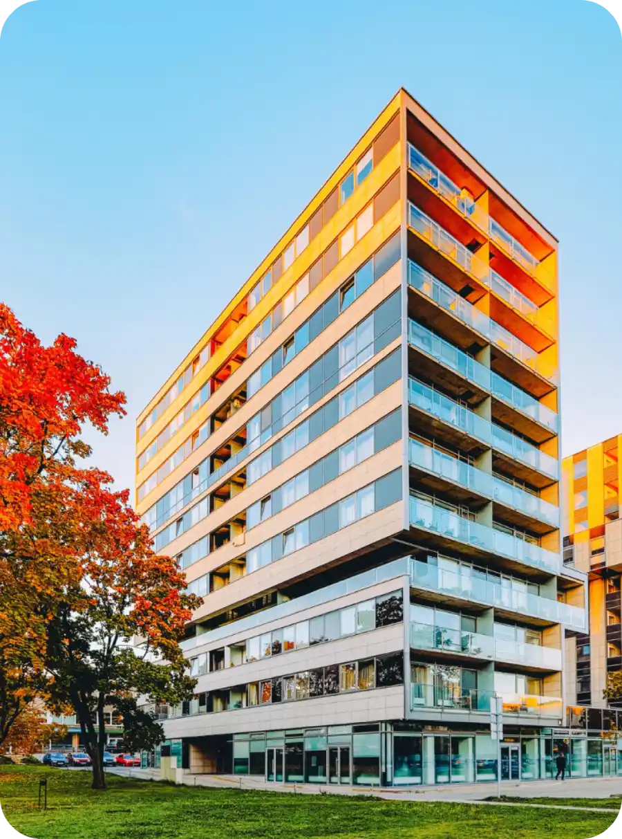 Edificio moderno con finestre a specchio, sole al tramonto, alberi con foglie autunnali, cielo sereno.