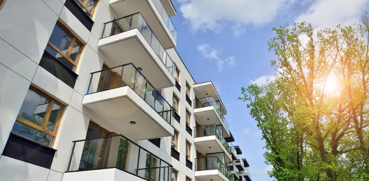 Moderni appartamenti con balconi in vetro, cielo blu e albero verde a lato durante il giorno.