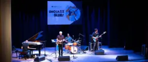 Gruppo jazz suona su un palco con pianoforte, sassofono, batteria e basso, palco illuminato.