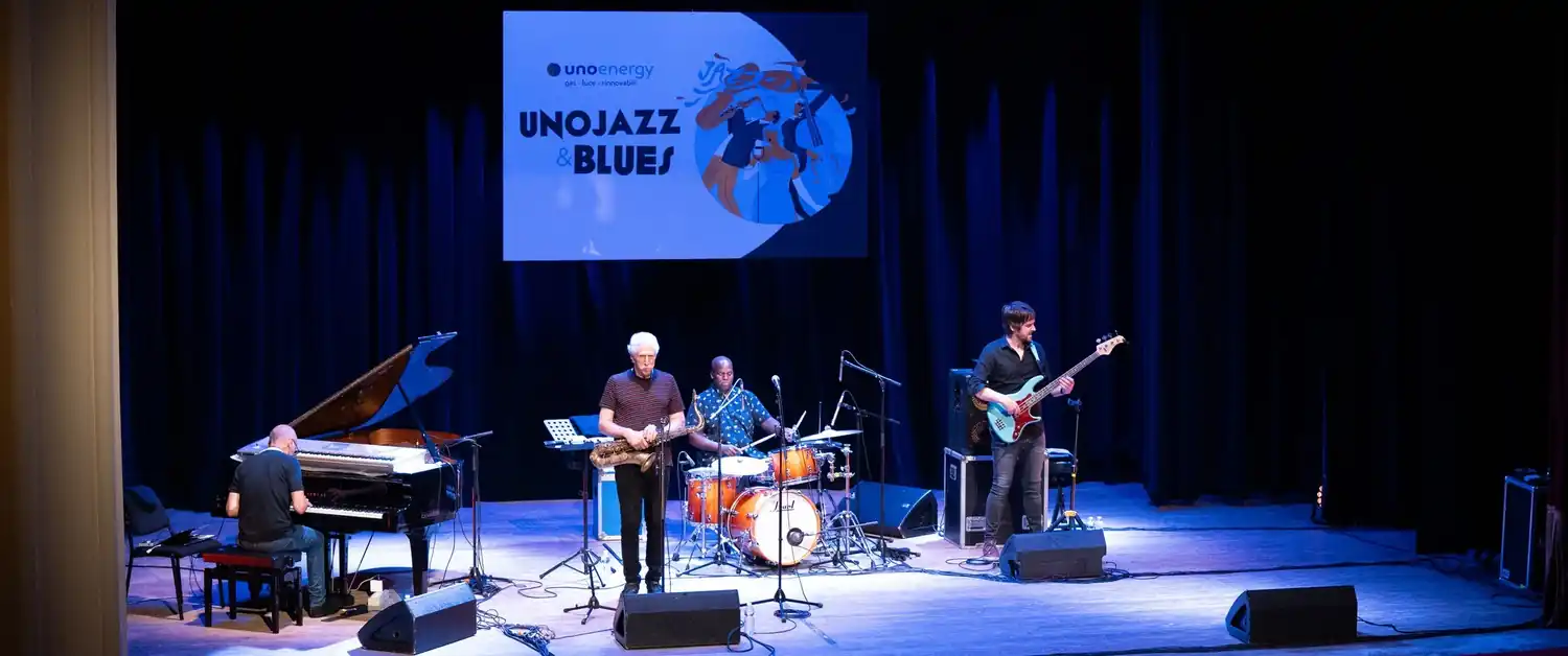 Gruppo jazz suona su un palco con pianoforte, sassofono, batteria e basso, palco illuminato.