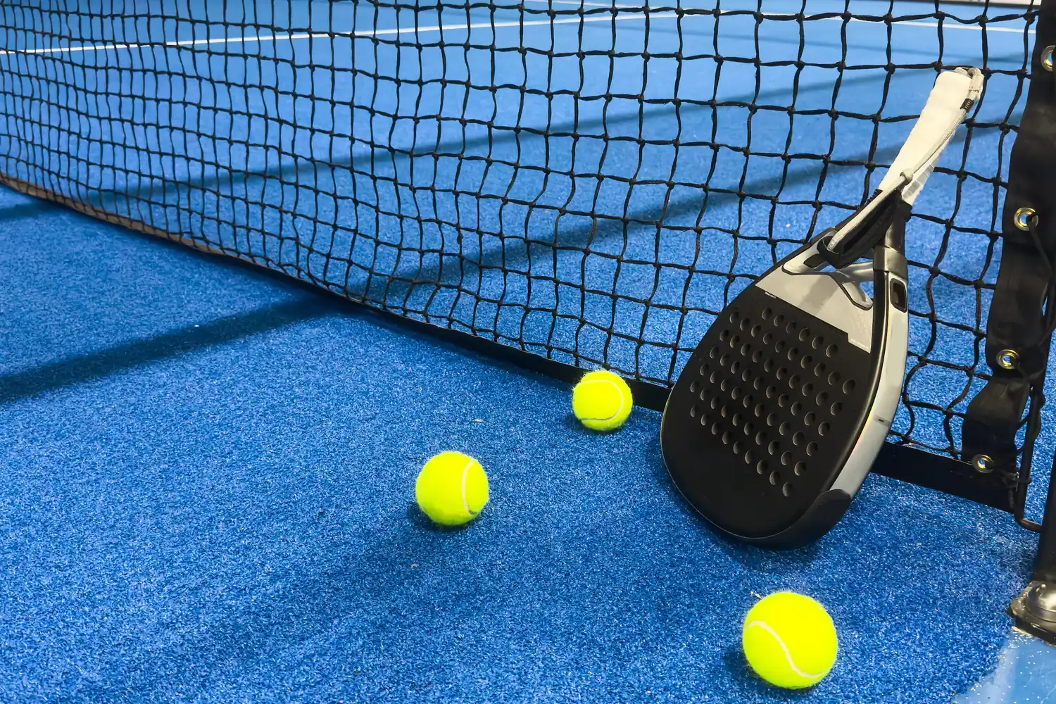 Racchetta padel e tre palline gialle su campo blu con rete nera.