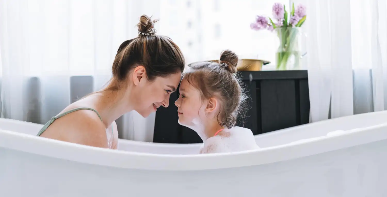 Madre e figlia sorridenti in vasca da bagno, fronte a fronte, con fiori sullo sfondo.
