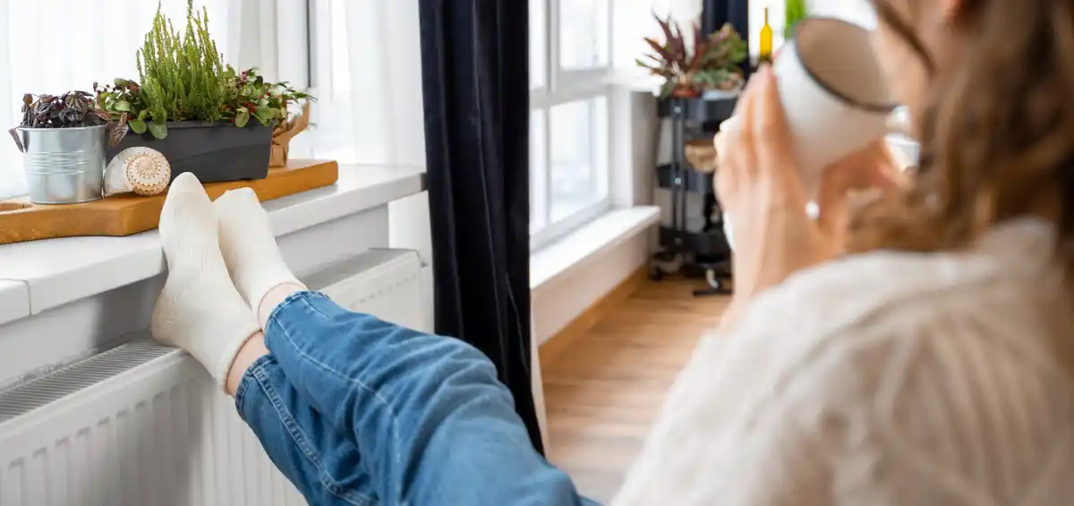Persona seduta, gambe su un termosifone, beve da una tazza accanto a piante su un davanzale.