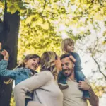 Famiglia sorridente all'aperto, due bambini sulle spalle dei genitori in un parco.