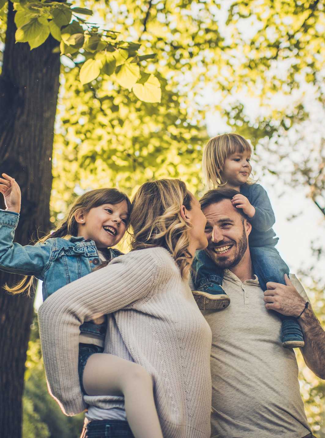 Famiglia felice all'aperto, madre e padre con due bambini piccoli su spalle tra gli alberi.