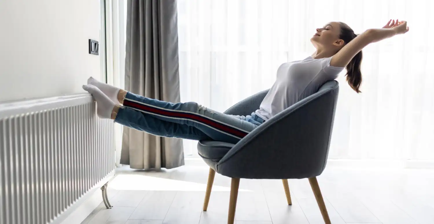 Donna seduta su poltrona, gambe appoggiate a termosifone, si stiracchia con soddisfazione, indossa calze bianche.
