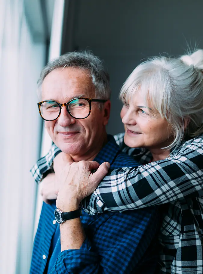Coppia anziana sorridente in posa abbracciata vicino a una finestra.