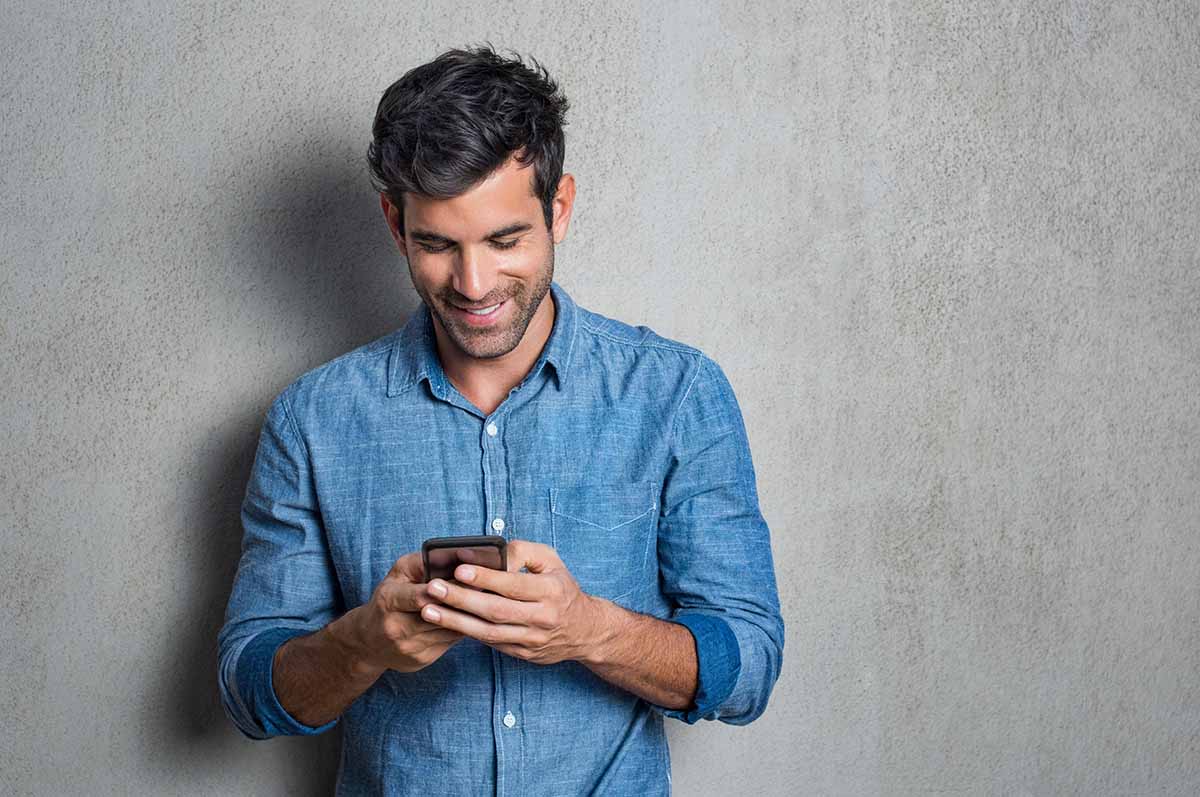 Uomo sorridente in camicia di jeans guarda smartphone contro parete grigia.