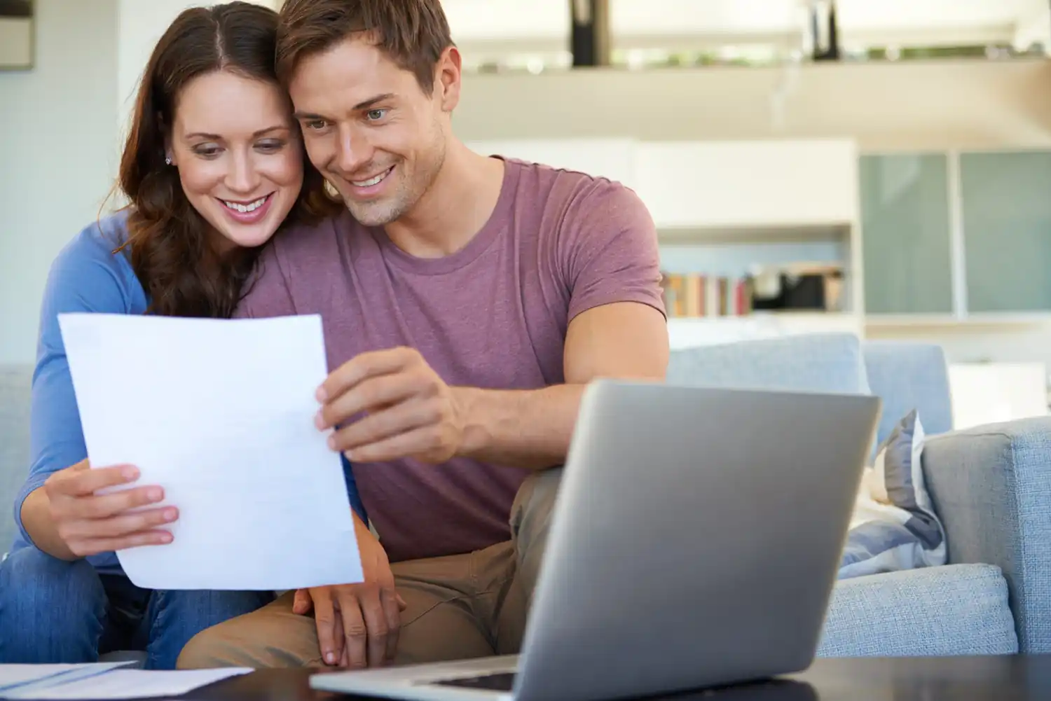 Coppia sorridente controlla fogli vicino a laptop in soggiorno moderno.