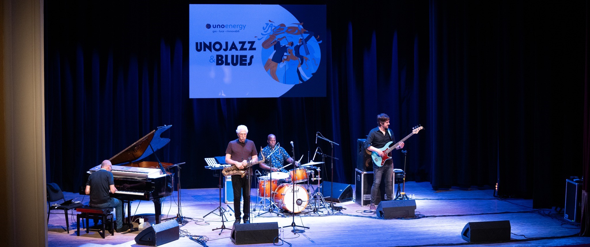 Gruppo jazz suona su un palco con pianoforte, sassofono, batteria e basso, palco illuminato.