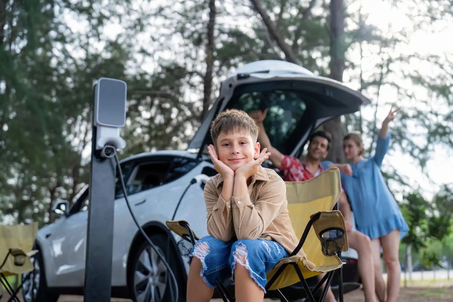 Bambino seduto su sedia da campeggio, con auto elettrica in carica sullo sfondo, famiglia felice in auto aperta.