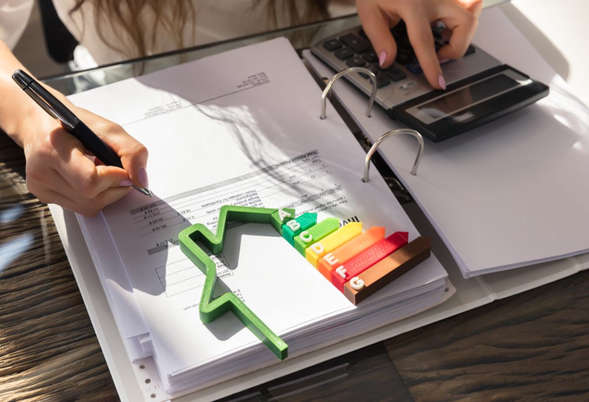 Persona scrive documenti, diagramma efficienza energetica, calcolatrice e raccoglitore su scrivania.