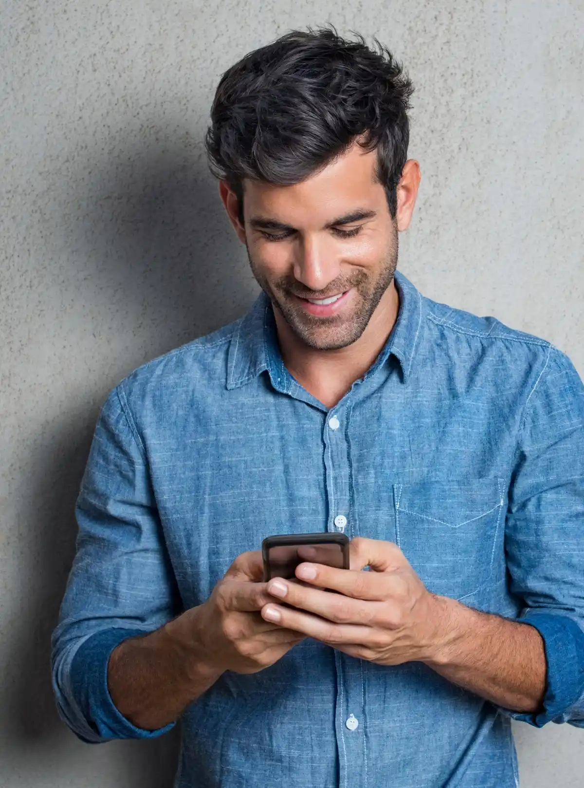 Uomo sorridente in camicia di jeans guarda smartphone contro parete grigia.