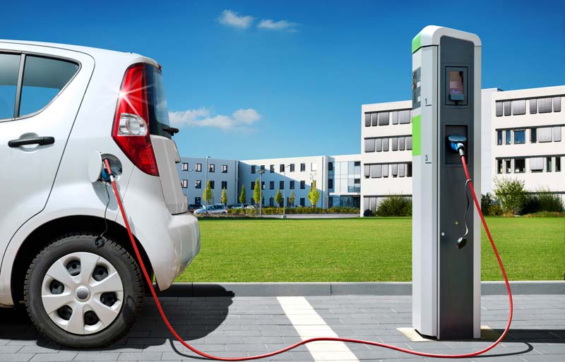 Auto elettrica in carica presso una stazione di ricarica pubblica in area urbana, sullo sfondo edifici moderni.