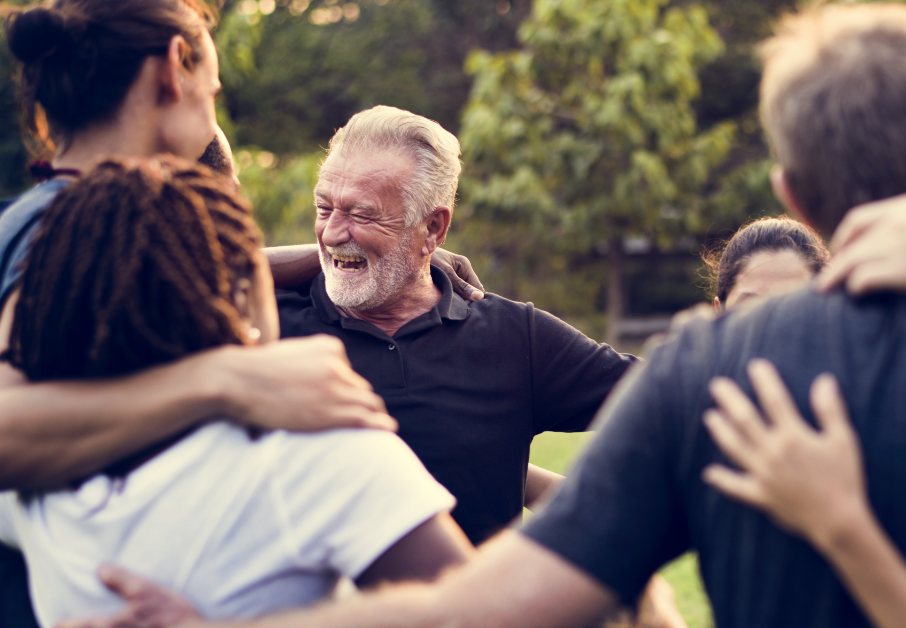 Gruppo di persone diverse abbracciate in cerchio all'aperto sorridendo, scena di amicizia e comunità.