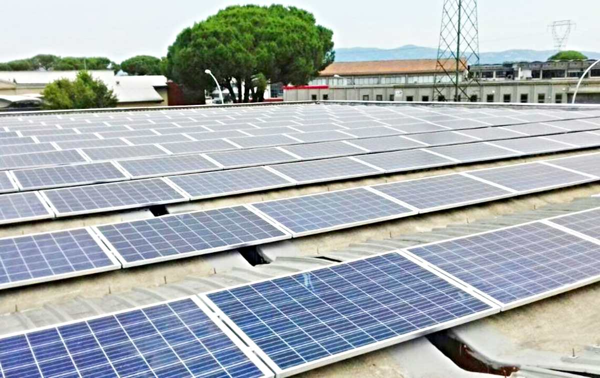 Pannelli solari su un tetto piatto in un'area industriale con alberi e strutture sullo sfondo.