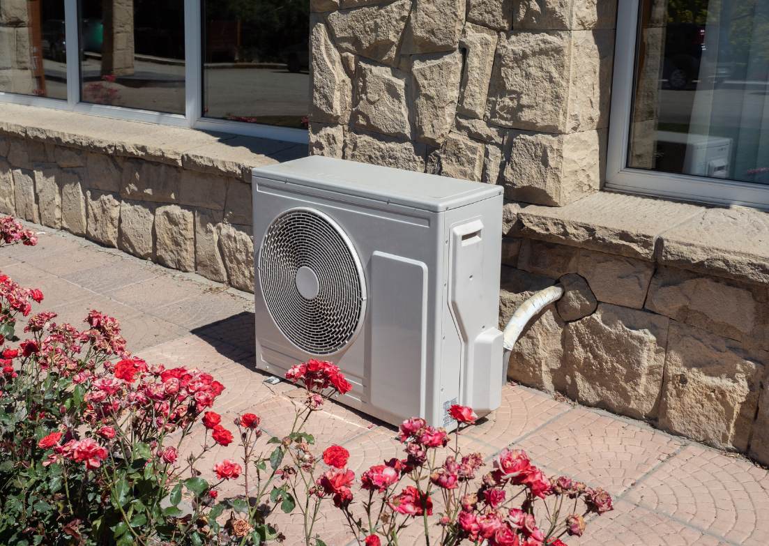 Unità esterna di climatizzazione accanto a un edificio in pietra, circondata da rose rosse fiorite.