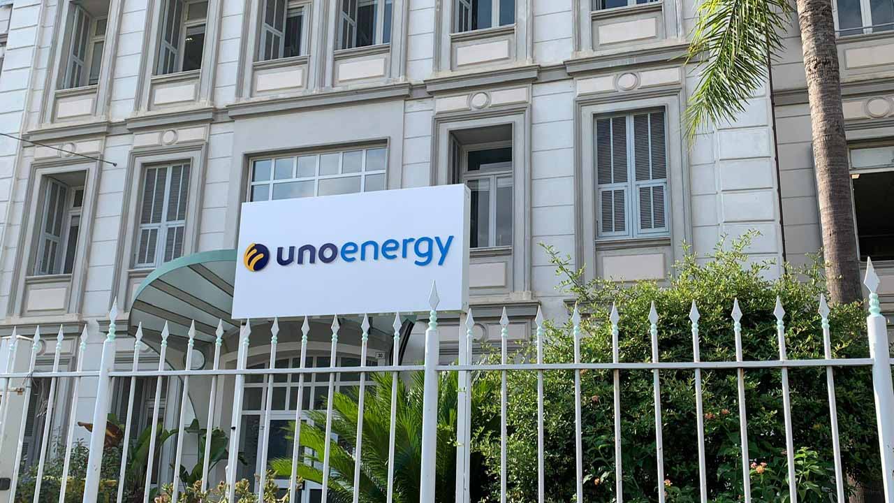 Insegna UnoEnergy su edificio con finestre e vegetazione in primo piano, recinzione bianca.