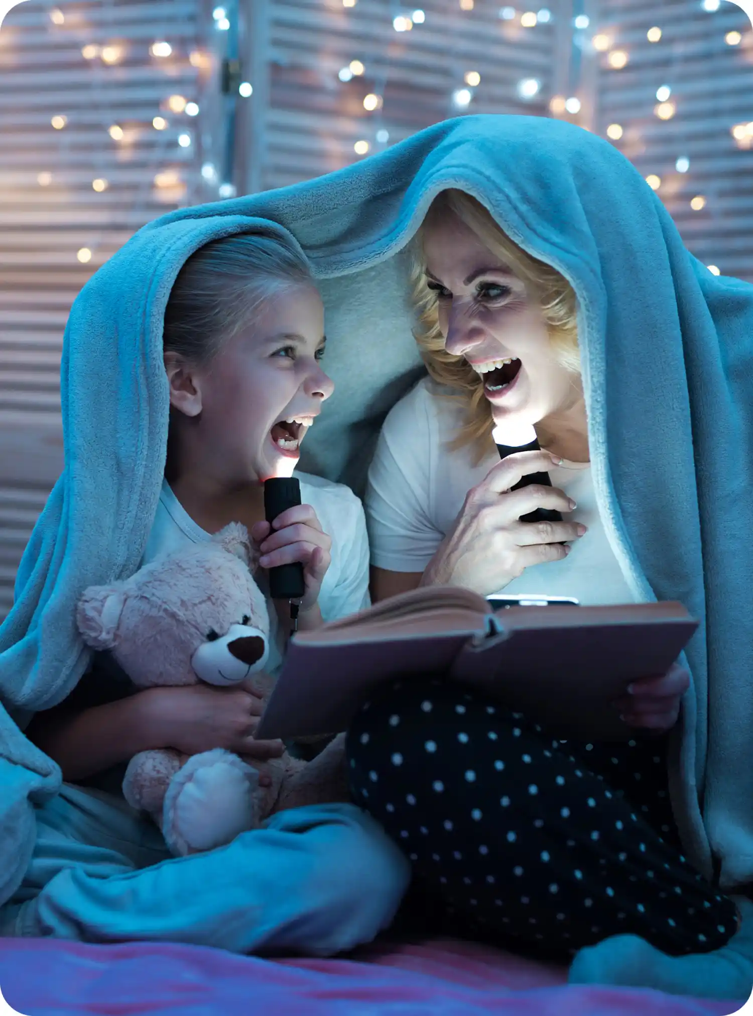 Bambina e adulto leggono un libro sotto una coperta con torce, luci soffuse creano un'atmosfera accogliente.