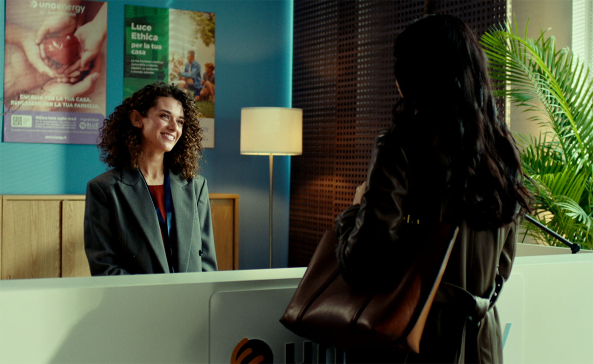 Donna sorridente alla reception in un ufficio, interagisce con una cliente di spalle. Poster e pianta sullo sfondo.