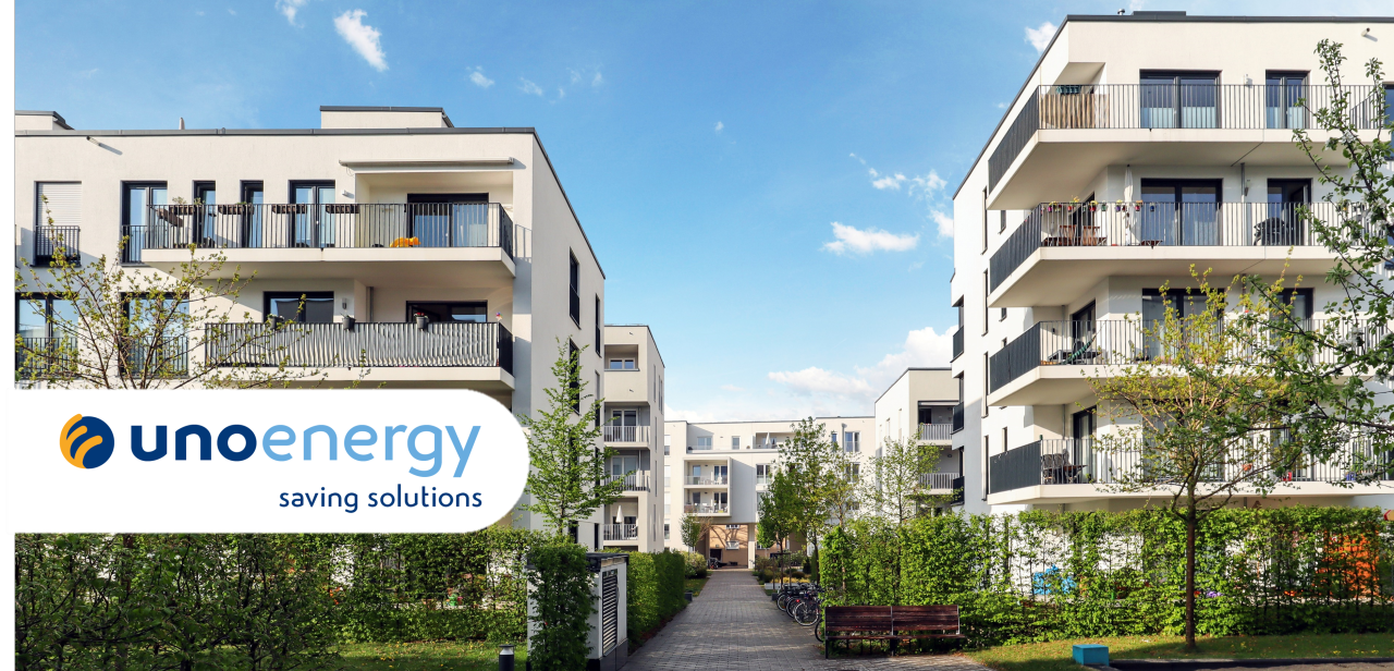 Edifici residenziali moderni con logo Unoenergy in basso a sinistra, cielo blu e verde in primo piano.