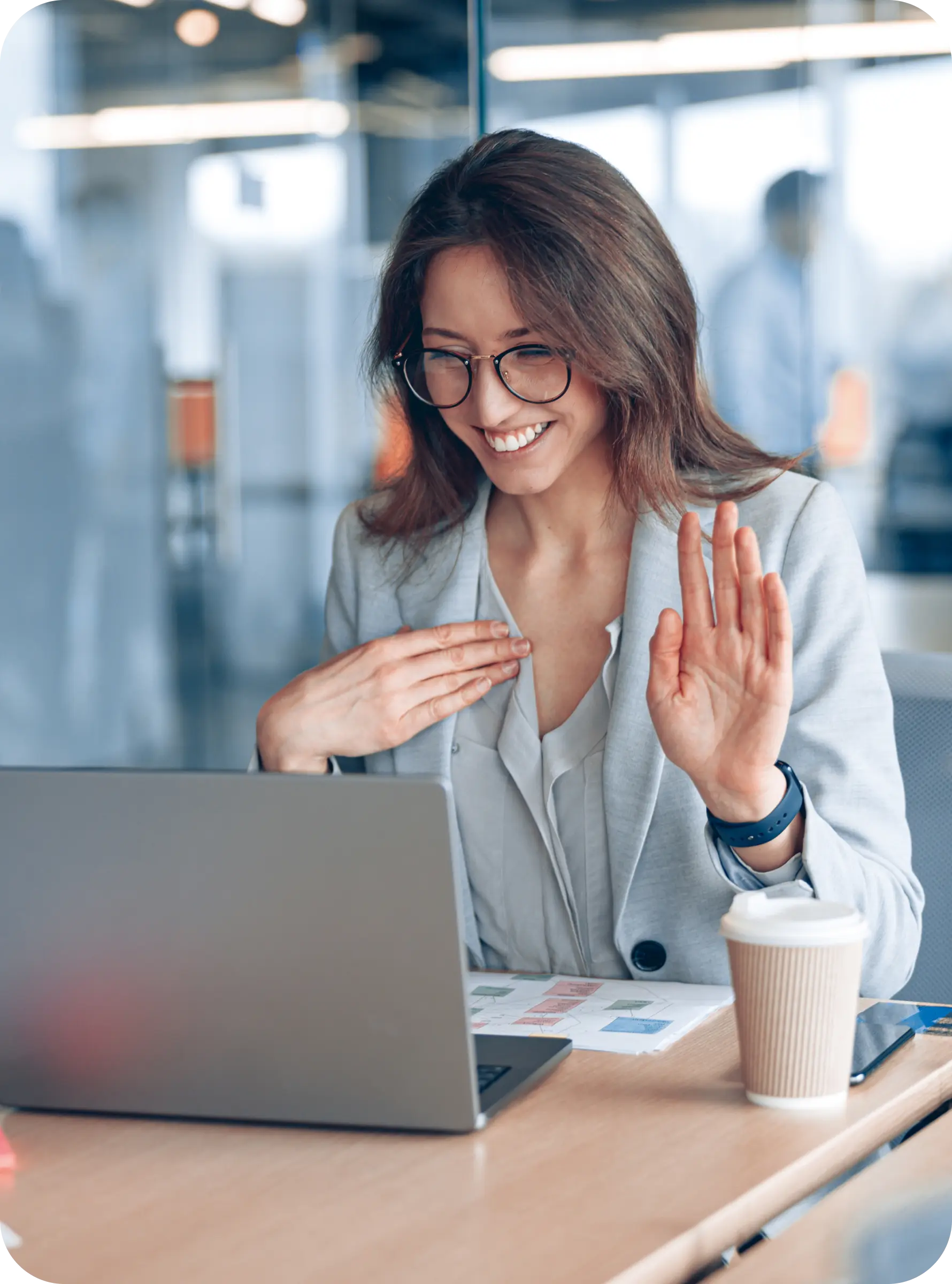 Donna sorridente in videochiamata al laptop in ufficio, con caffè su tavolo.
