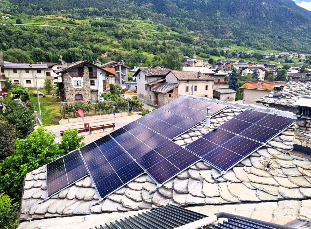 Pannelli solari su tetto di casa in pietra con villaggio e colline verdi sullo sfondo.