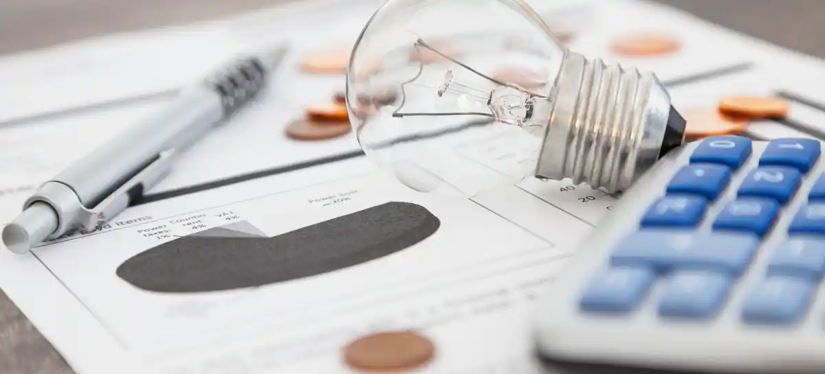 Lampadina, calcolatrice e penna su bolletta energetica con monete sparse.
