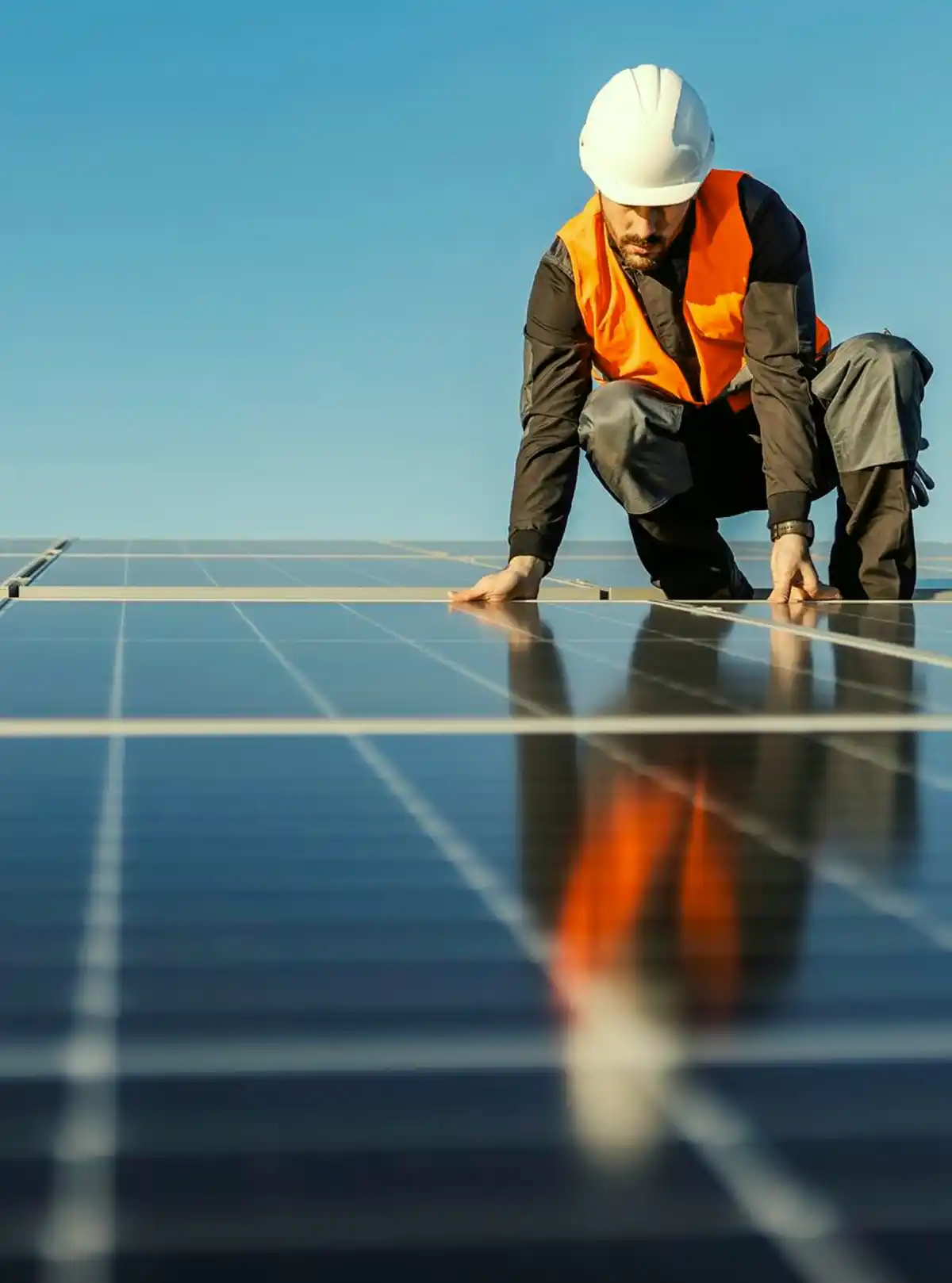 Tecnico ispeziona pannelli solari su tetto con giubbotto arancione e casco protettivo bianco.