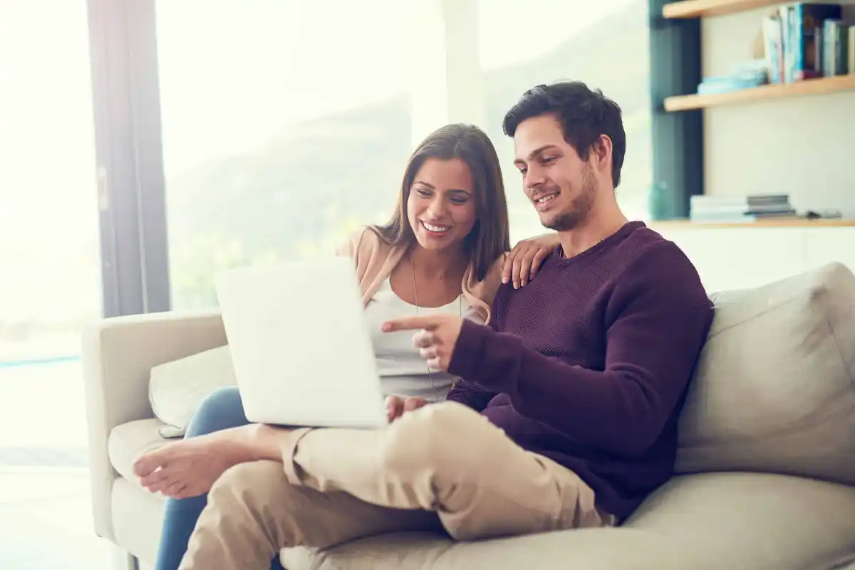 Coppia sorridente seduta sul divano mentre guarda un laptop in una stanza luminosa.