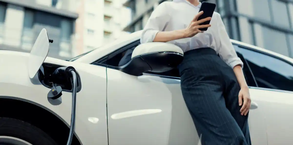 Donna appoggiata a un'auto elettrica in carica con smartphone in mano, città sullo sfondo.