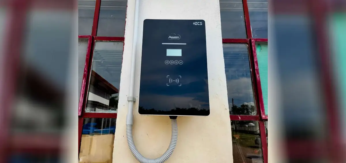 Caricabatterie industriale nero montato a parete, con display e pulsanti, davanti a una finestra con telaio rosso.