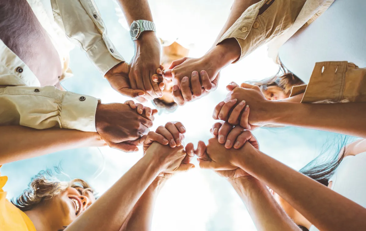 Gruppo di persone in cerchio si tengono per mano, cielo azzurro sullo sfondo.