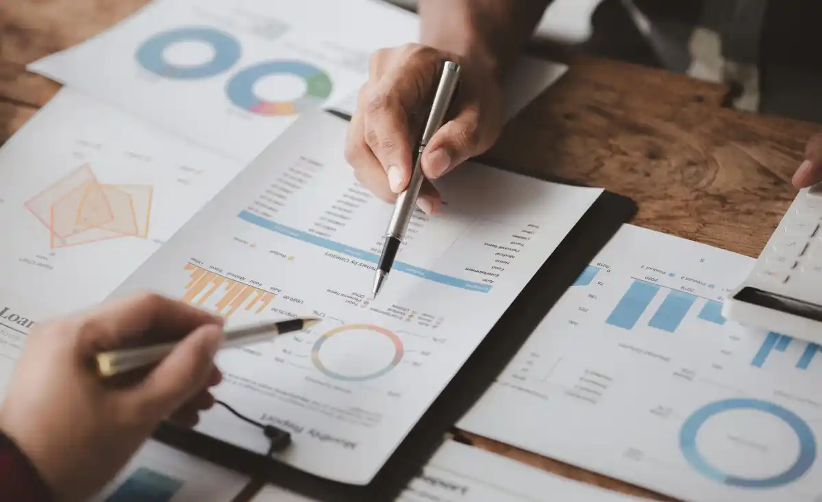Persone analizzano dati finanziari su grafici e tabelle, utilizzando penne e calcolatrice, su tavolo di lavoro.
