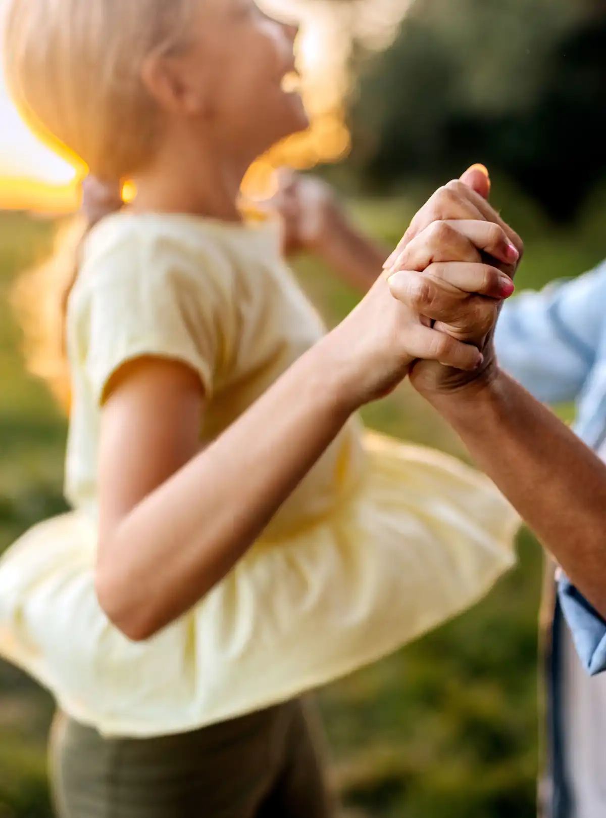 Bambina in abito giallo balla all'aperto tenendo le mani di un adulto al tramonto.