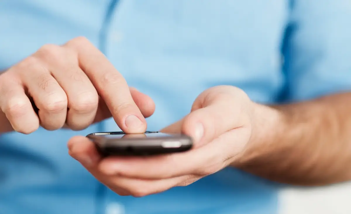 Persona usa smartphone, toccando lo schermo con dito indice, indossa camicia azzurra a maniche lunghe.